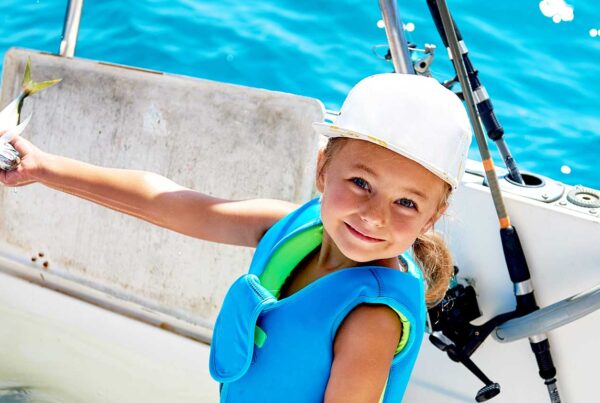 Photo of a little blond girl with blue eyes wearing a blue life vest and white hat, holding a fish she had just caught while on a motorboat.