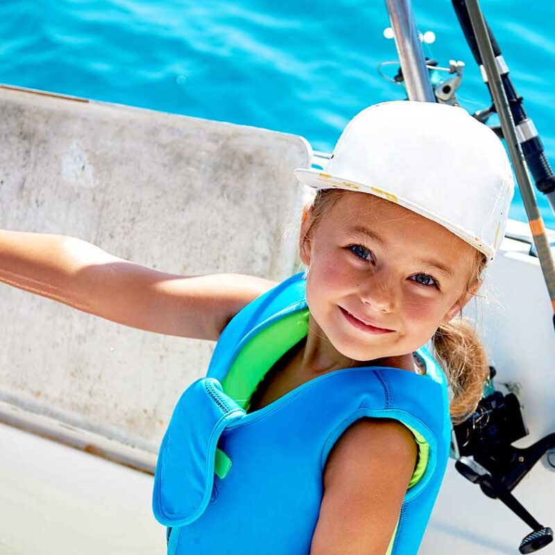 Photo of a little blond girl with blue eyes wearing a blue life vest and white hat, holding a fish she had just caught while on a motorboat.